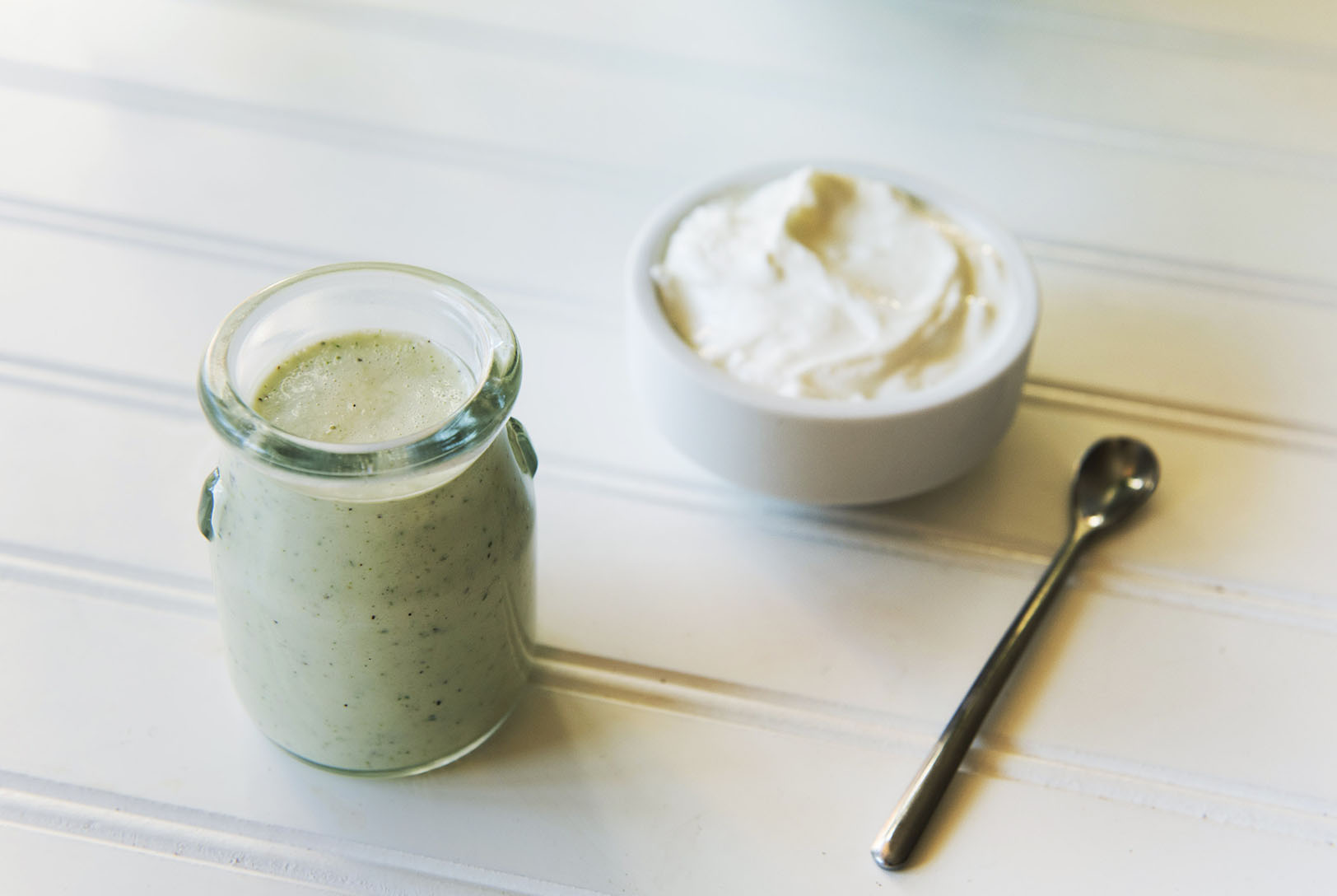 Creamy green Tzatziki Dressing in a small glass jar next to a bowl of plain yogurt and a spoon on a white wooden surface.