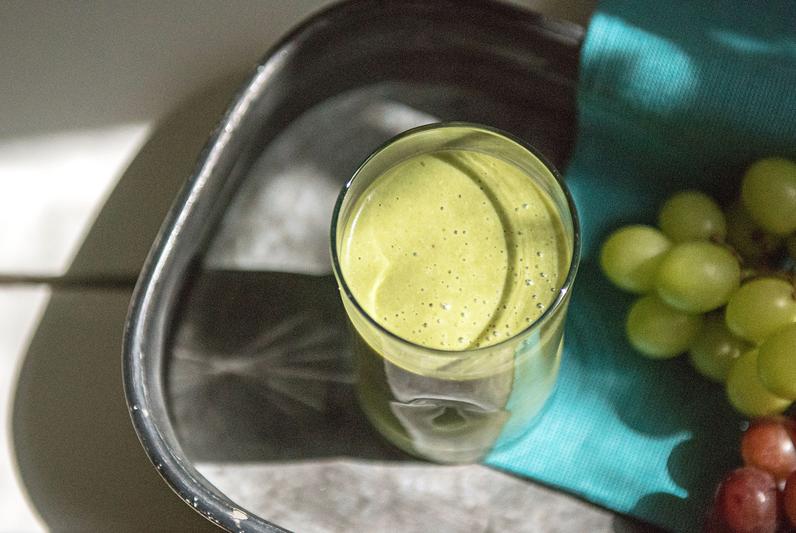 green smoothie in a tall glass on a tray next to fresh green and purple grapes