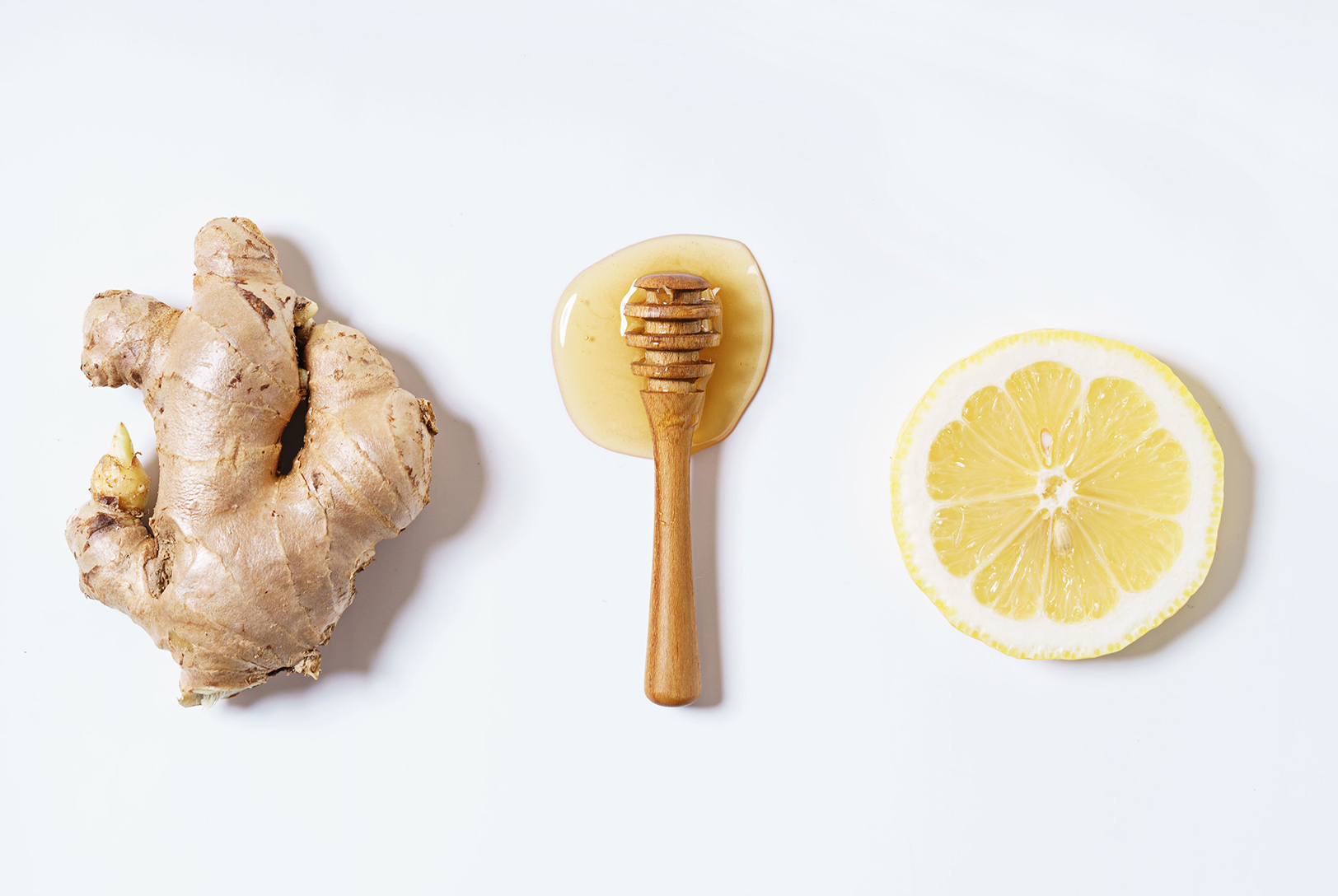 A whole piece of ginger root, a wooden honey dipper with honey, and a slice of fresh lemon on a clean white surface.
