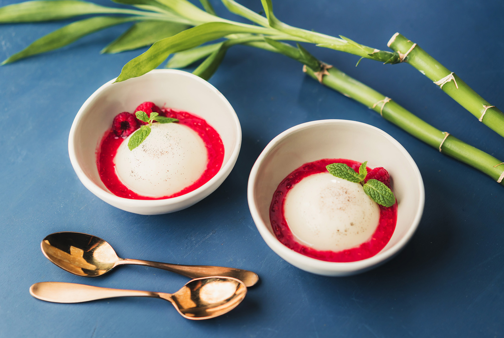 Two white bowls containing smooth, white Almond Panna Cotta with a vibrant red raspberry sauce, garnished with raspberries and mint.