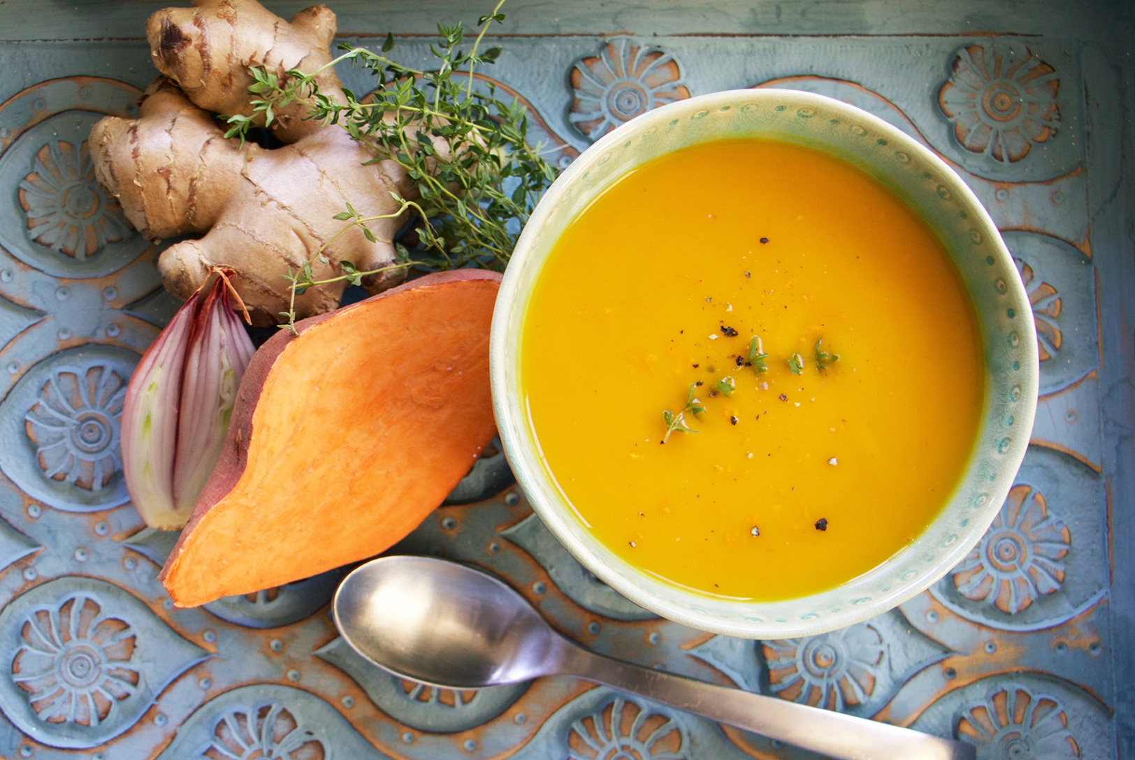 Overhead view of creamy Sweet Potato Bisque in a light green bowl with ginger, shallot, and herbs on a decorative blue surface.