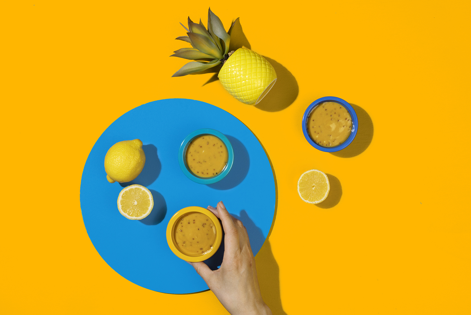 Overhead shot of Honey Mustard Dressing in colorful bowls, surrounded by fresh lemons and a pineapple decoration on a yellow surface.