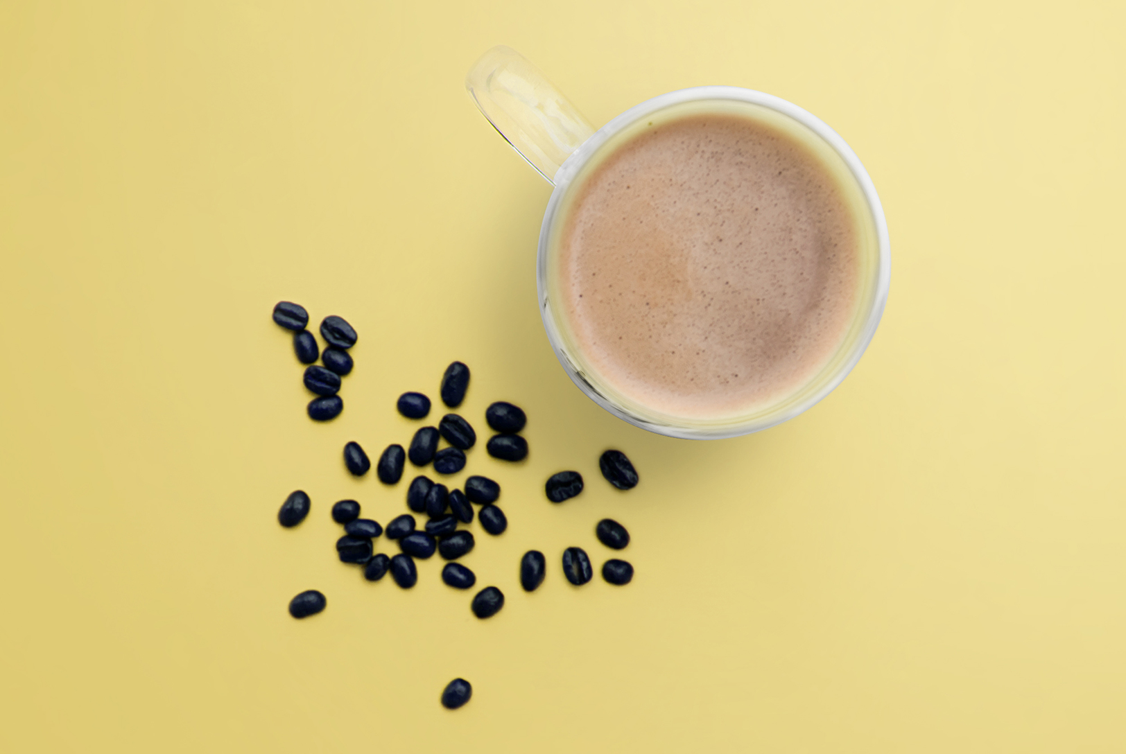 A clear glass mug filled with Creamy Coffee Smoothie, viewed from above, with a scattering of coffee beans on a yellow background.