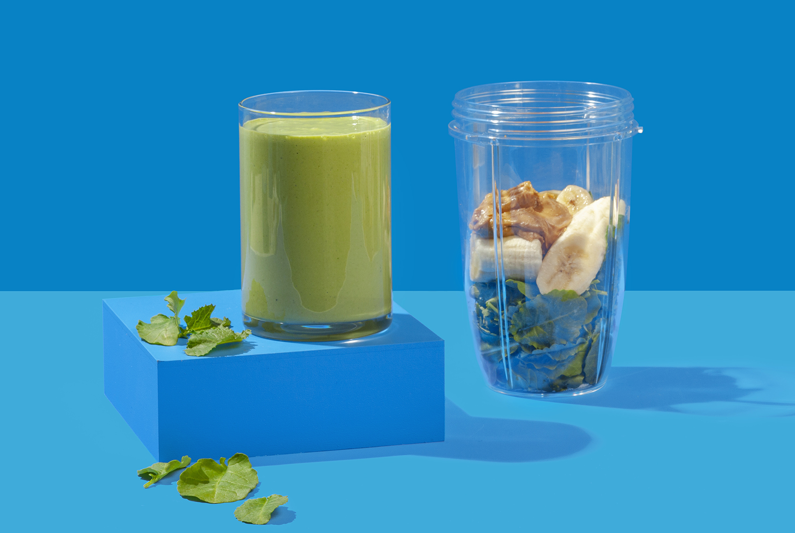 Breakfast Smoothie in a glass next to a blender cup filled with spinach, banana, and peanut butter on a vibrant blue background.