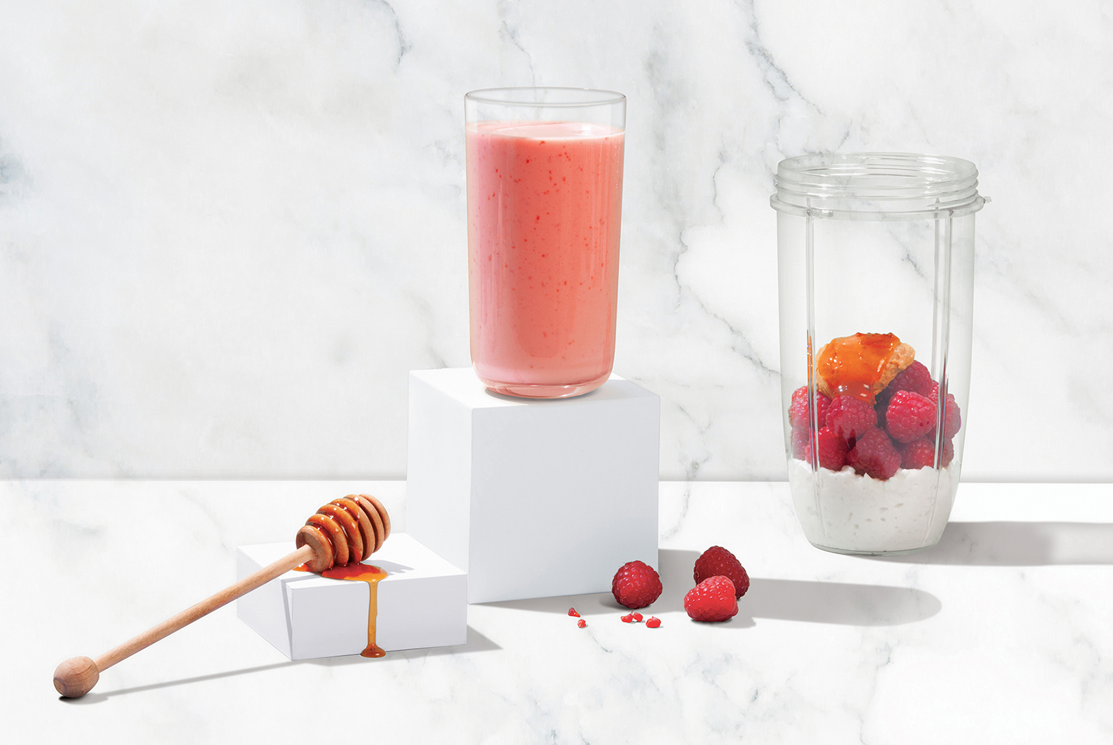 Raspberry Protein Smoothie in a glass with ingredients, nearby a blender cup with cottage cheese, raspberries, peanut butter and honey.