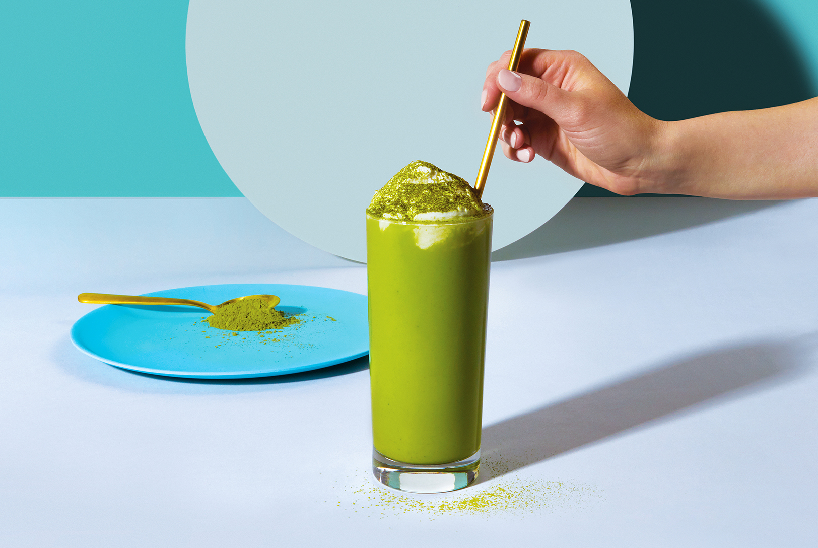 A tall glass of green Matcha Frappe topped with whipped cream and matcha powder, with a gold straw being held by a hand.