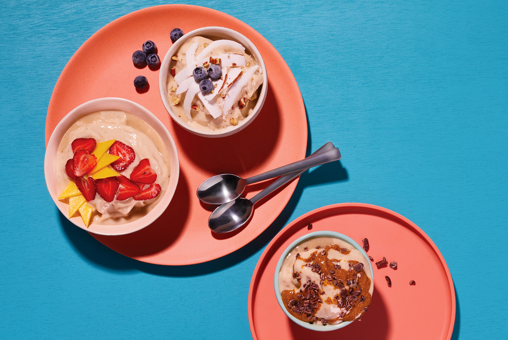 Three bowls of Nice Cream, with different toppings like strawberries, mango, blueberries, coconut flakes, and chocolate drizzle, arranged on coral plates.