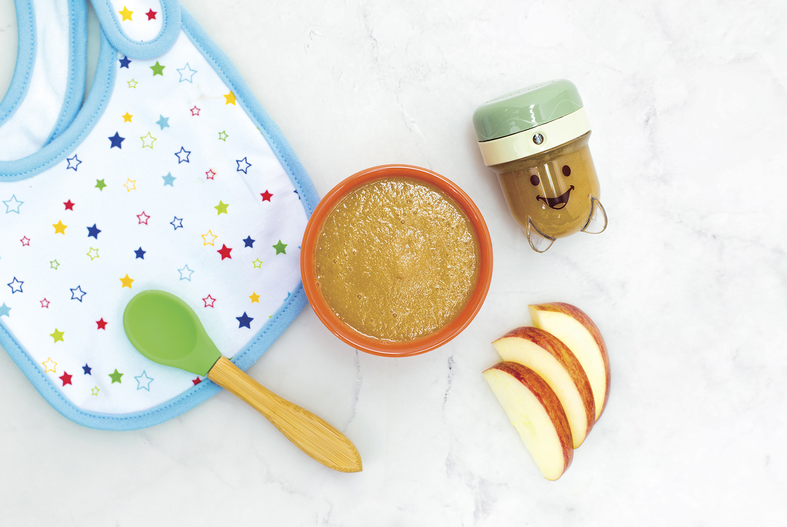 A bowl of Apple Purée next to slices of fresh apple, a baby spoon, and a small baby food maker with a baby bib in the corner.