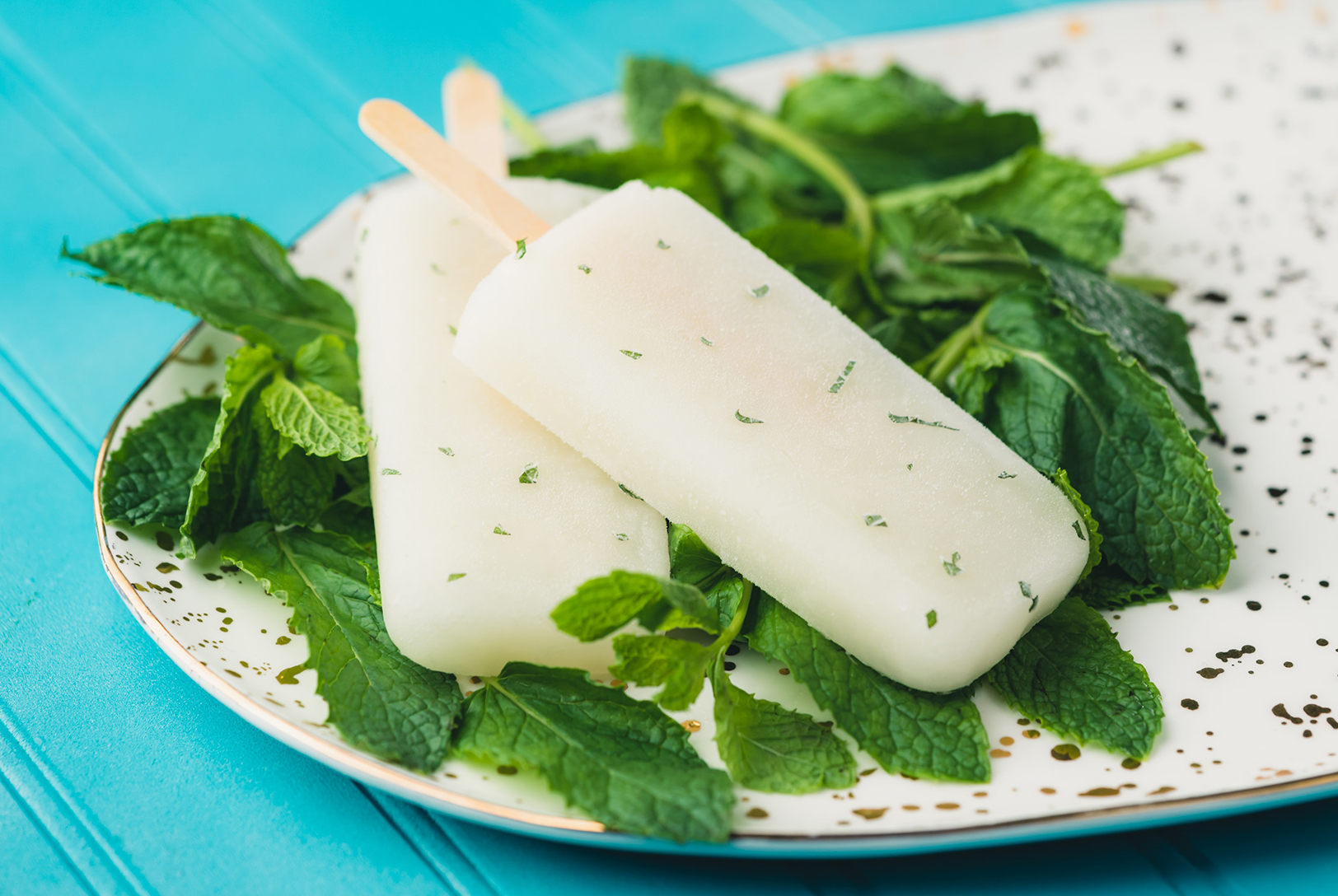 Two Green Tea Peppermint Popsicles with wooden sticks, on fresh mint leaves on a white plate with gold speckles.