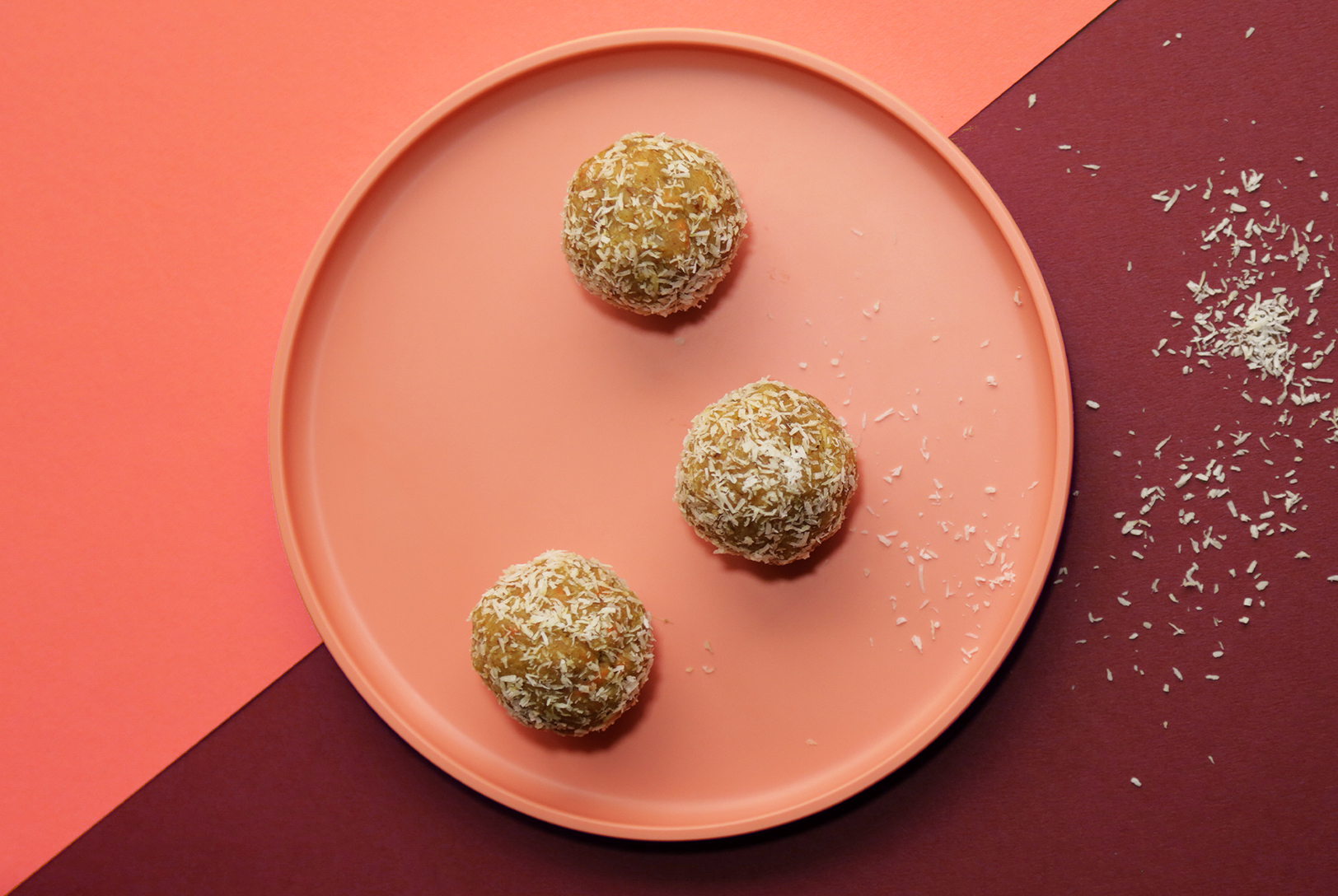 Three round Carrot Cake Bites, coated in shredded coconut, arranged on a plate against a diagonally divided background of light orange and dark red.