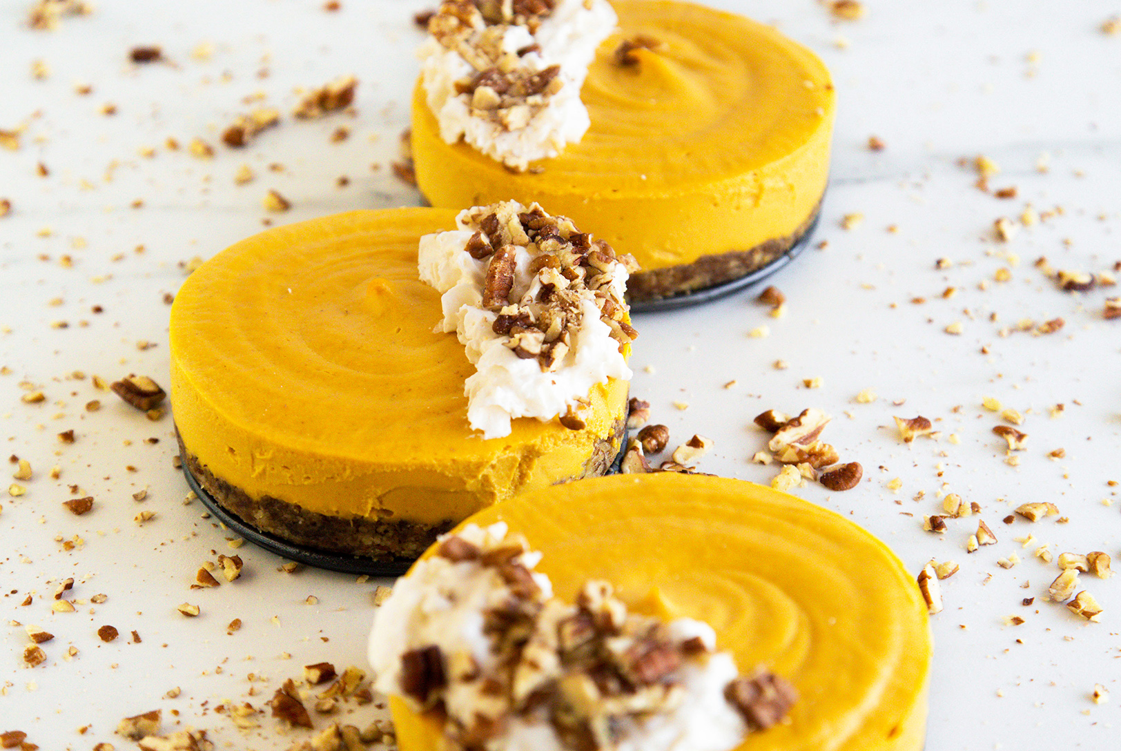 Three no-bake pumpkin cheesecakes topped with whipped cream and walnuts, surrounded by crushed walnuts.