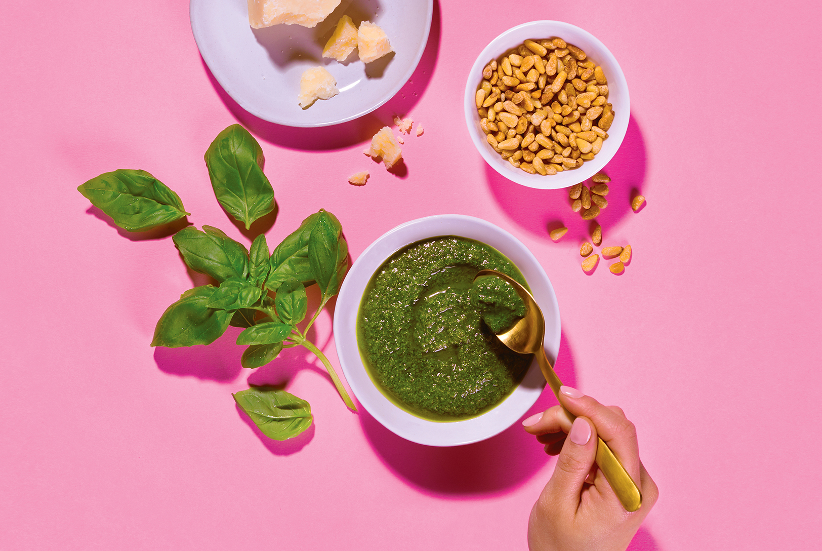 A bowl of Perfect Pesto with a spoon, basil leaves, a bowl of pine nuts, and Parmesan cheese on a bright pink background.