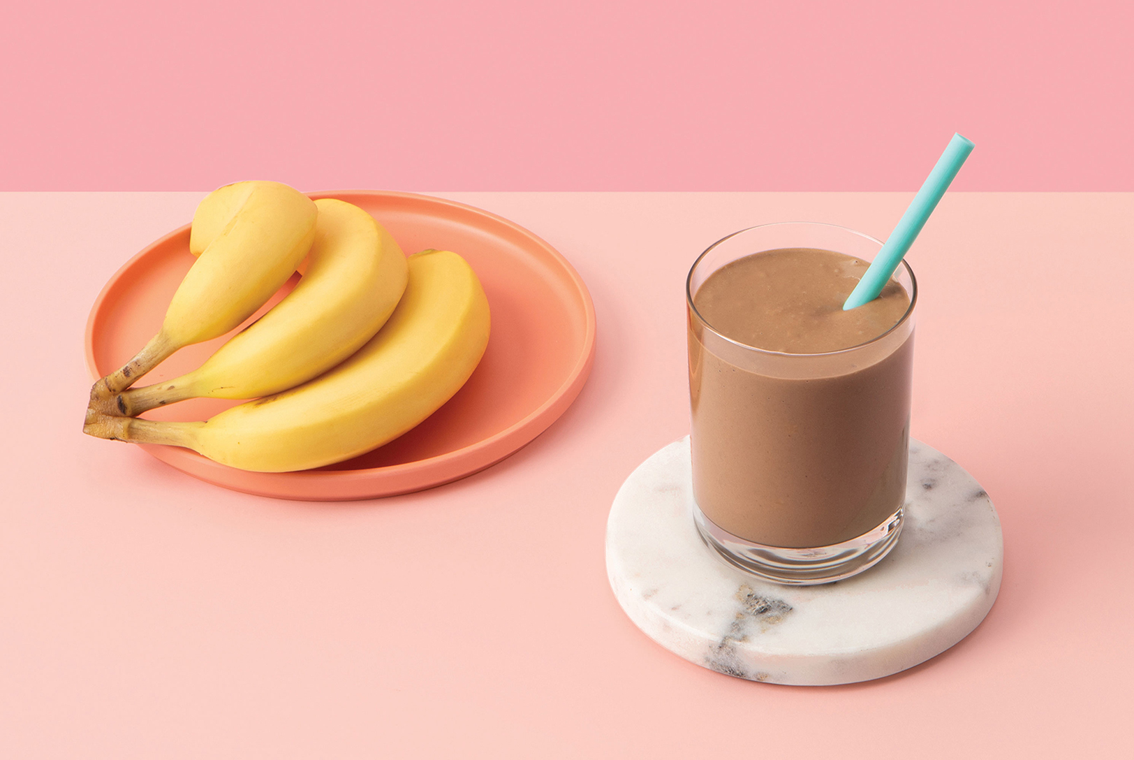 Nutrition Rich Smoothie in a glass with a blue straw next to a plate of ripe bananas on a soft pink background.