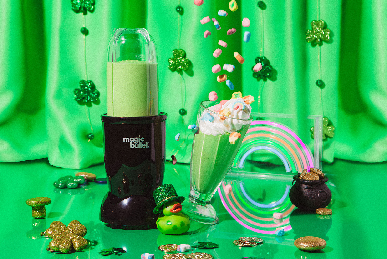 A magic bullet® next to a green Lucky Shamrock Smoothie in a glass with whipped cream and lucky charms, surrounded by St. Patrick's Day decorations.
