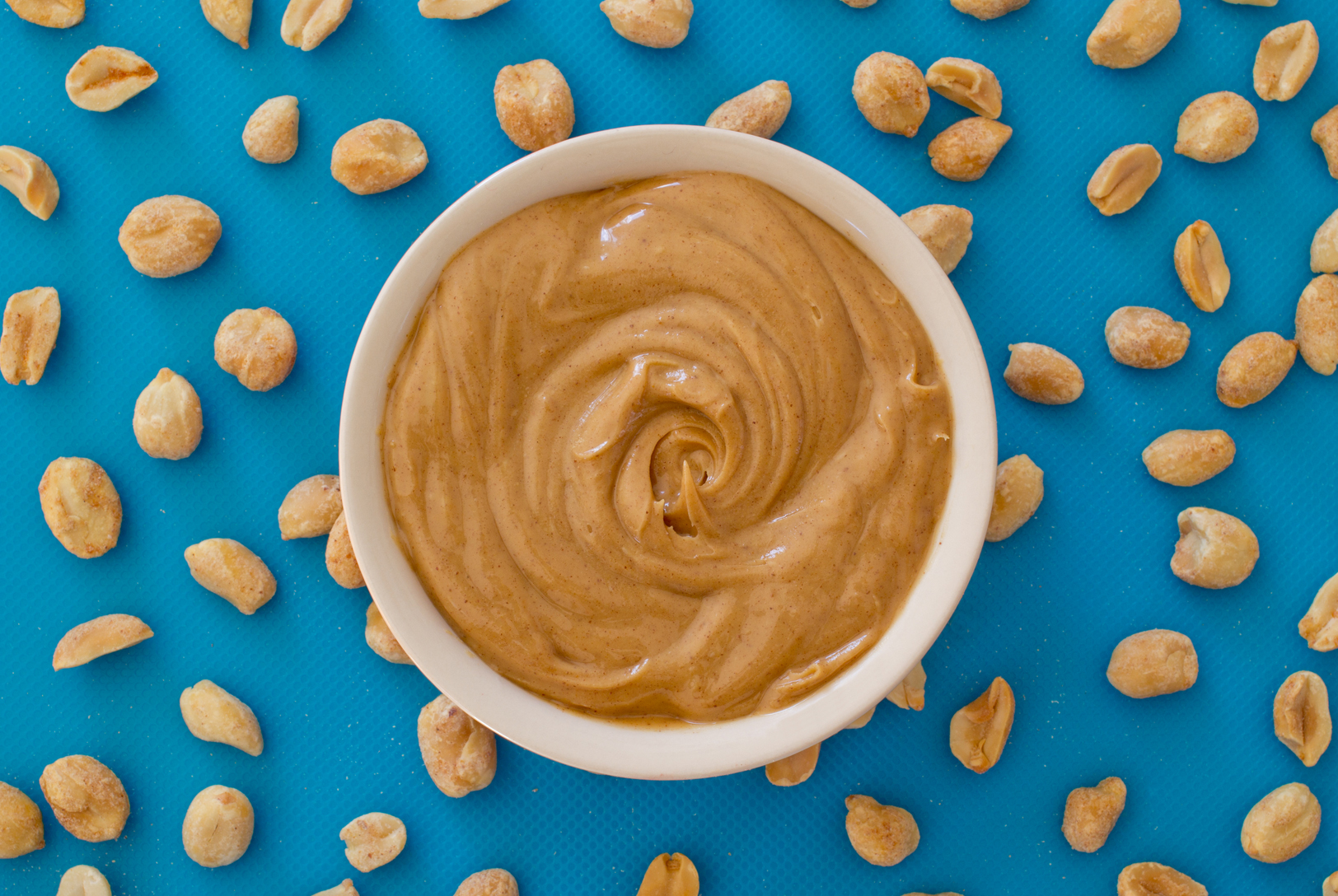 Creamy Peanut Butter in a white bowl, surrounded by scattered peanuts on a bright blue background.