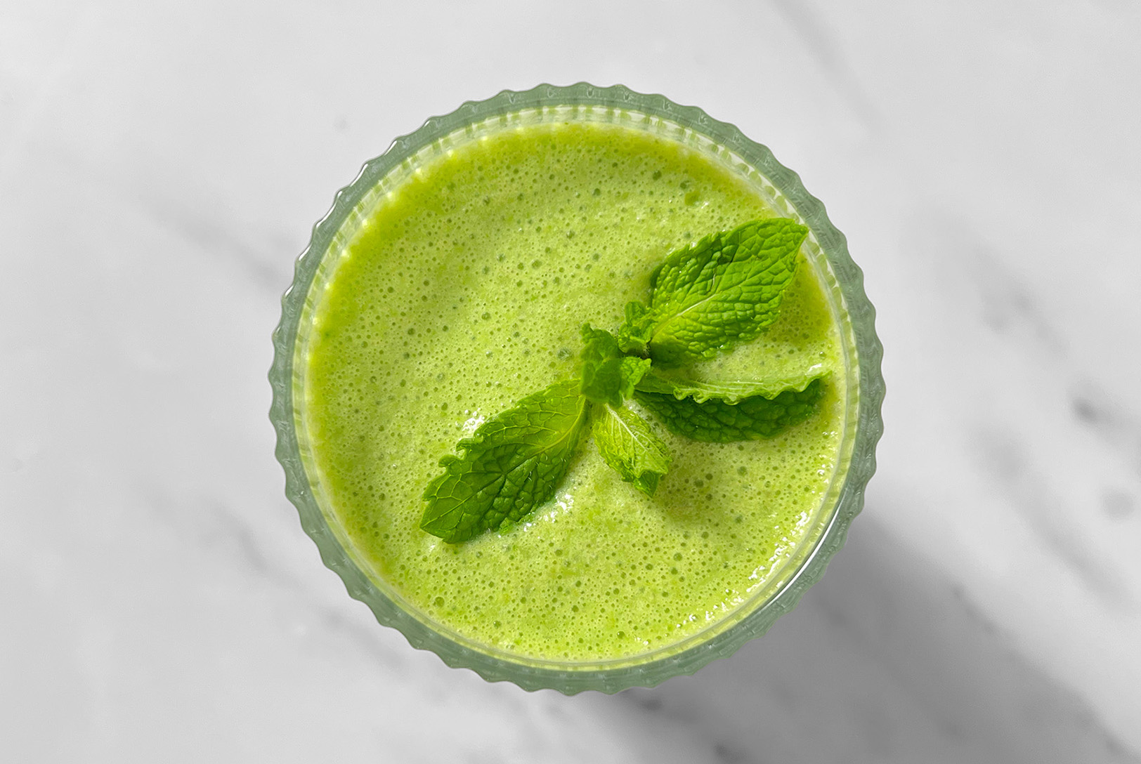 A hand holding a clear blender cup filled with spinach, sliced strawberries, and sliced bananas against a solid pink background.
