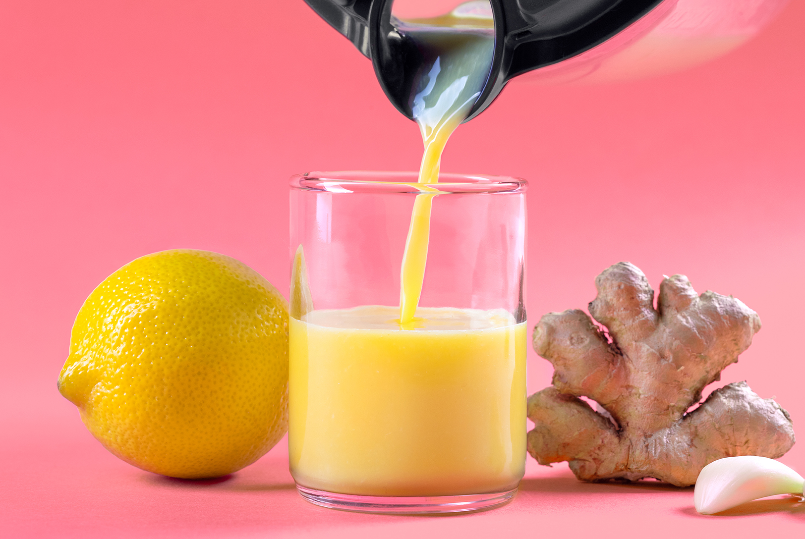 A Funky Fresh Ginger Shot is being poured into a glass, accompanied by fresh ginger, a whole lemon, and a garlic clove on a bright pink background.
