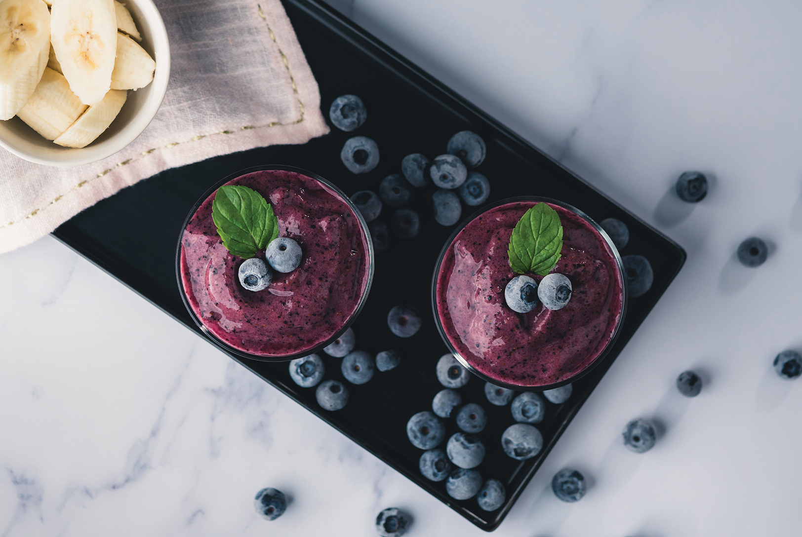 two blueberry smoothies in cups with fresh blueberries and banana slices on a black cutting board
