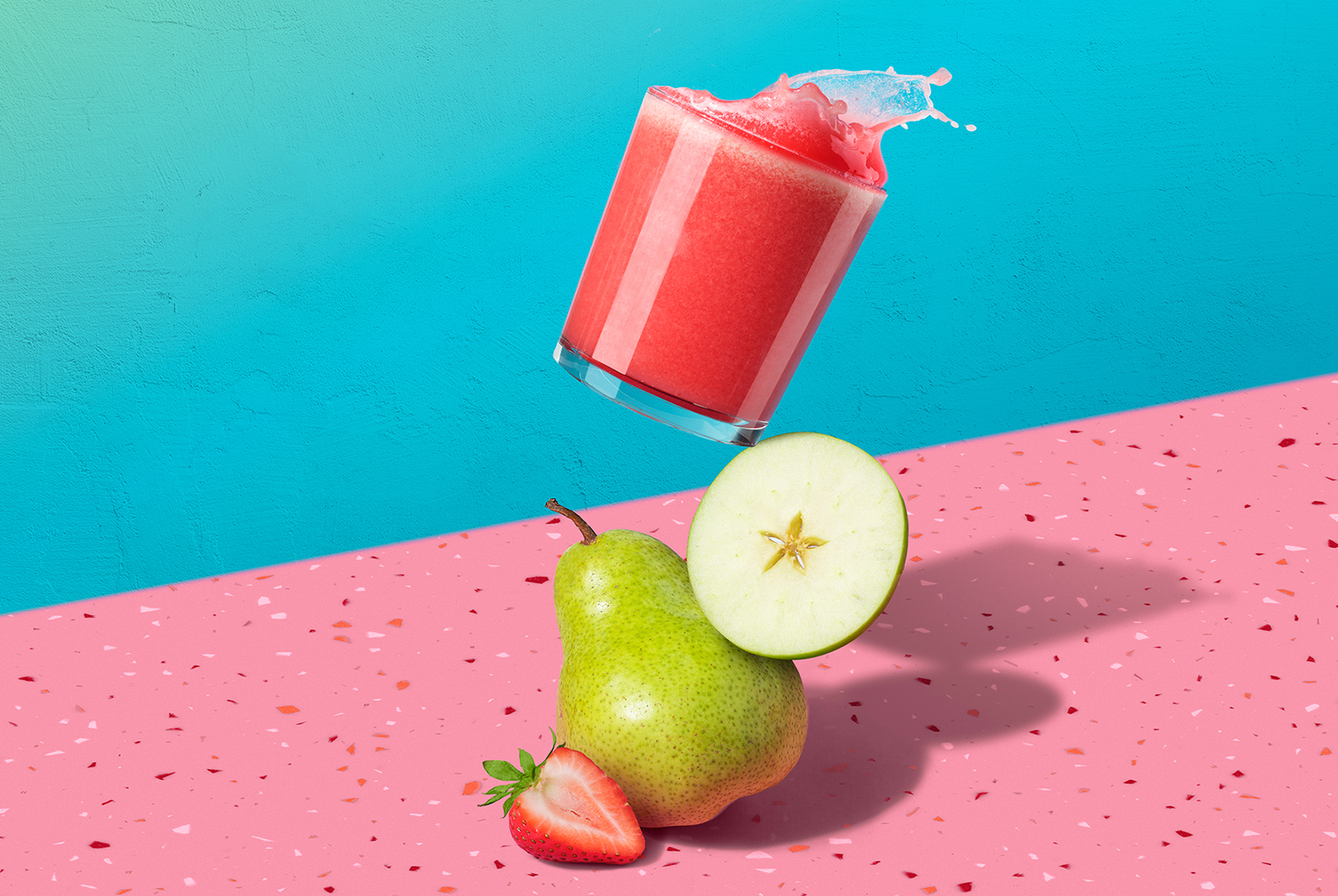 tilted glass sitting on top of pear with strawberry, apple, pear juice splashing on a pink counter with blue background