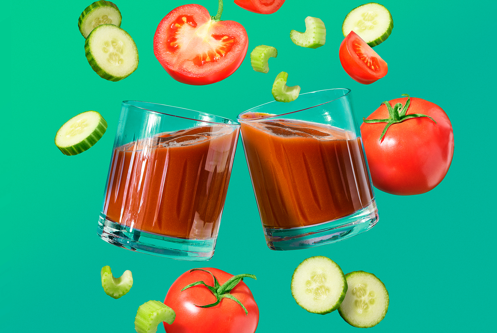 two glasses of tomato juice with tomatoes, cucumber and celery floating in front of green background