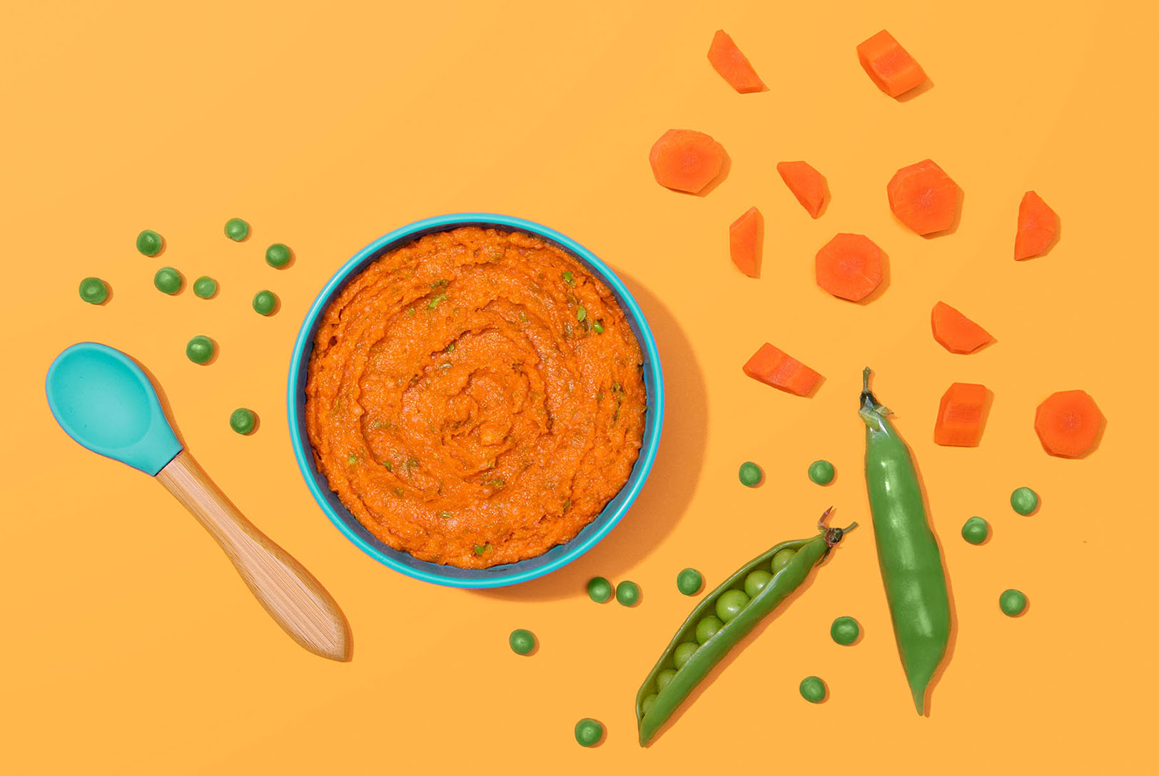 Pureed carrots in a blue bowl with pea pods and chopped carrots outside