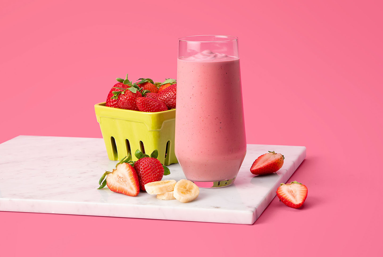 Pink strawberry banana smoothie in a glass on a cutting board with sliced fresh strawberries and bananas