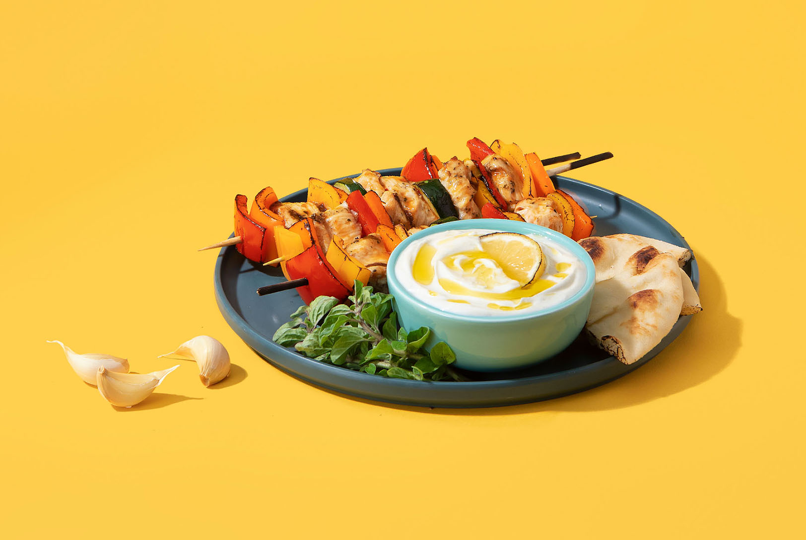 Bowl of homemade Garlic Spread (toum) plated alongside vegetable skewers, pita bread, herbs, and garlic with yellow background