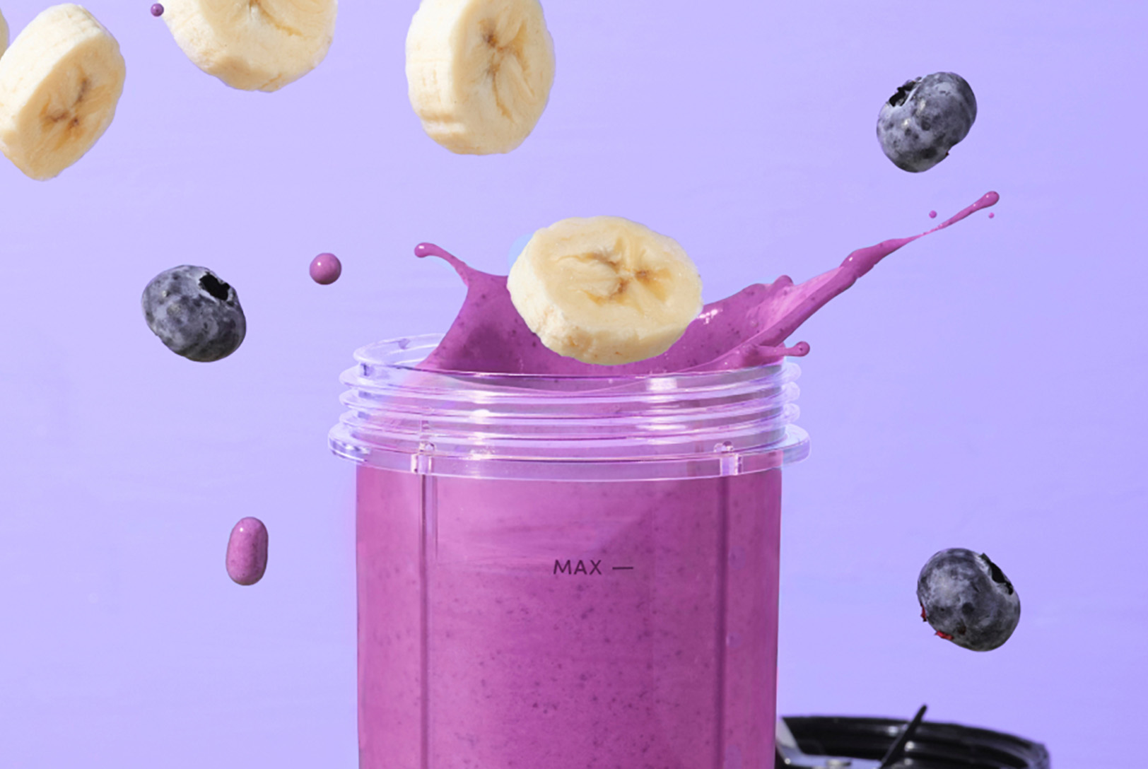 A blender cup with Berry Protein Smoothie with a splash, as sliced bananas and blueberries are dropped into it against a purple background.