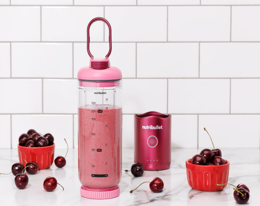 Pink nutribullet flex blender filled with a cherry smoothie, surrounded by fresh cherries in red bowls on a marble counter with white tile background.