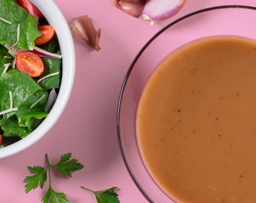 Bowl of green salad with tomatoes and bowl of brown salad dressing on pink surface.