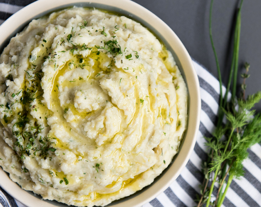 Bowl of mashed potatoes topped with oil drizzle and herbs on striped surface with green herbs.