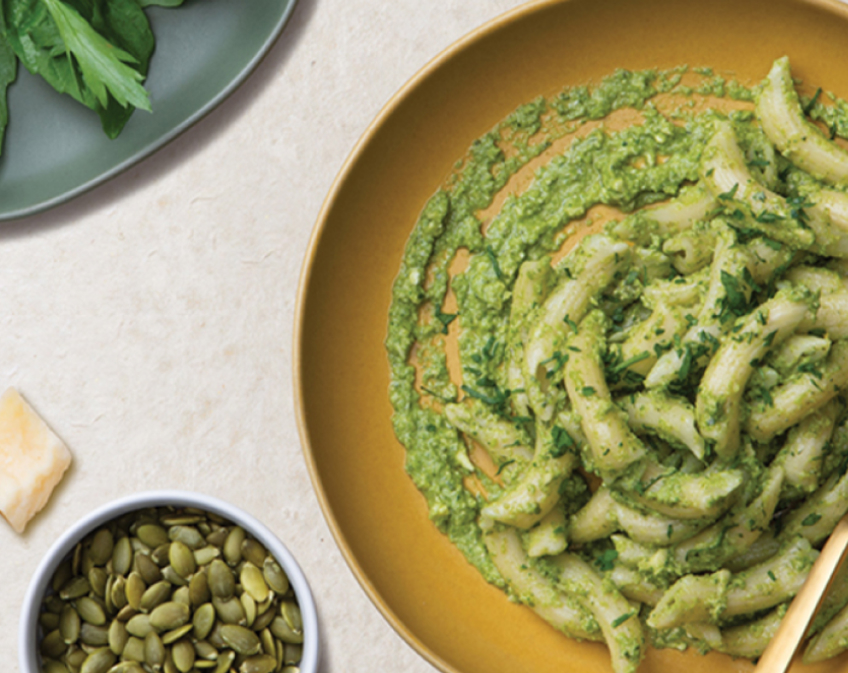 Yellow plate of pasta and green pesto sauce with herbs on white surface.