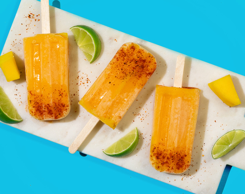 Three orange pospsicles with orange, lemon and mango slices against a blue and light tan background