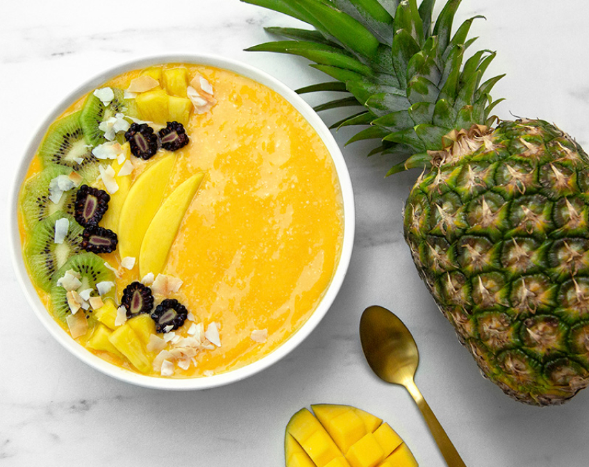 Overview of a white bowl filled with orange smoothie and topped with fruit next to a spoon, mango and a pineapple