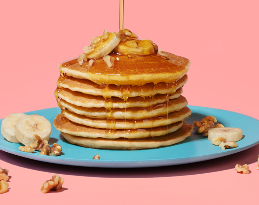 Syrup pouring on stack of pancakes topped with bananas and nuts on blue plate with pink background.