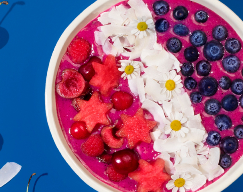 Purple acaí bowl topped with star shaped melon, berries, coconut flakes, flowers on blue background.