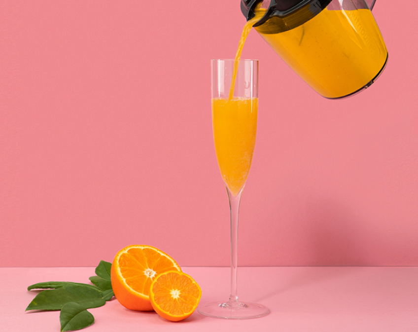 Pitcher pouring orange juice into champagne flute next to sliced orange and green leaves on pink background.