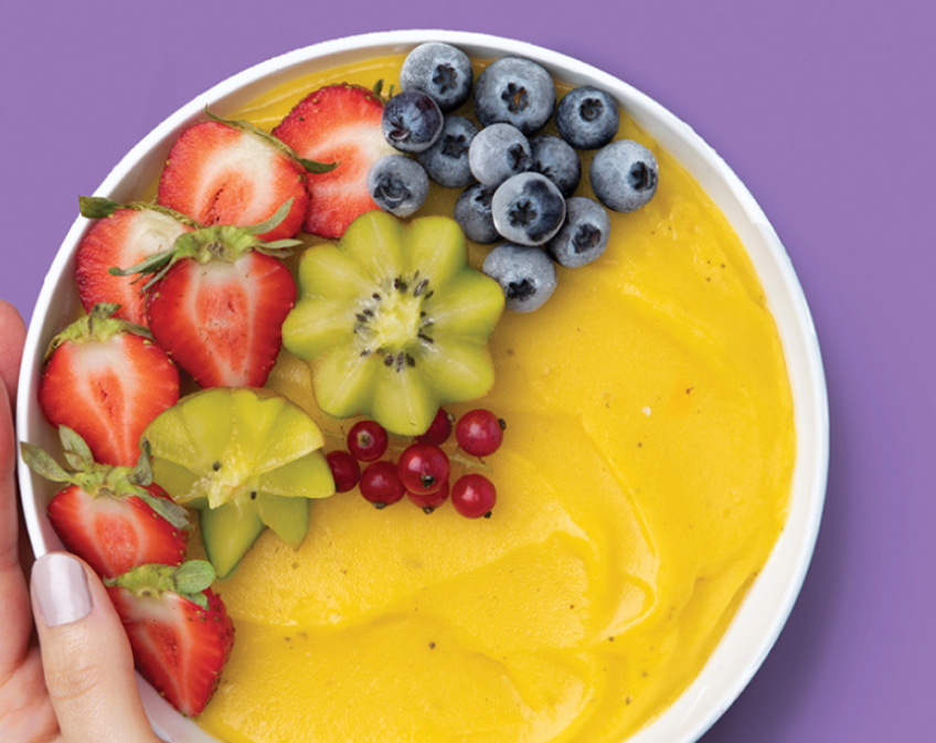 White bowl of yellow smoothie topped with strawberries, kiwis, blueberries on purple surface.