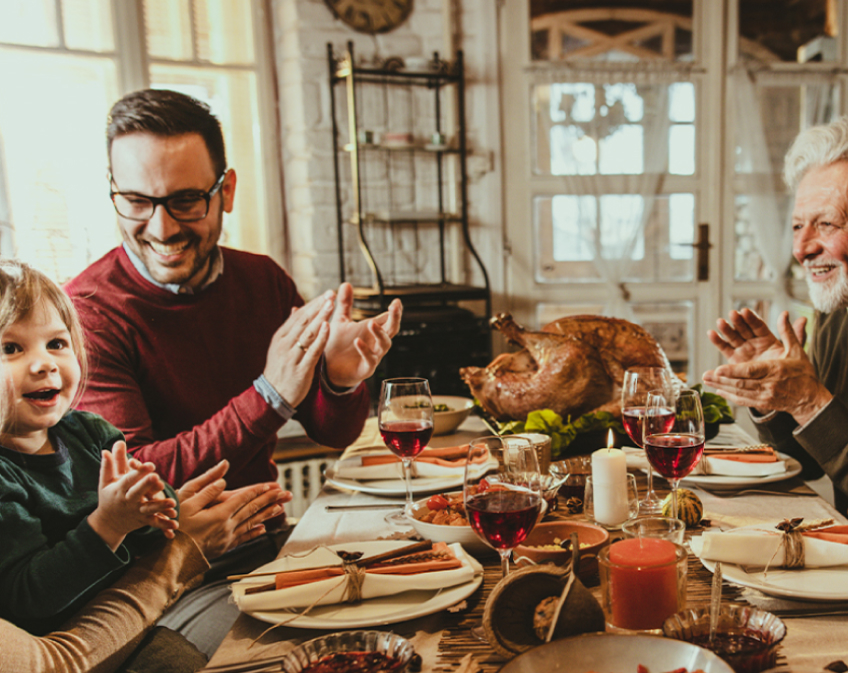 Scene of Thanksgiving dinner table with table settings, glasses of wine, turkey and smiling family.