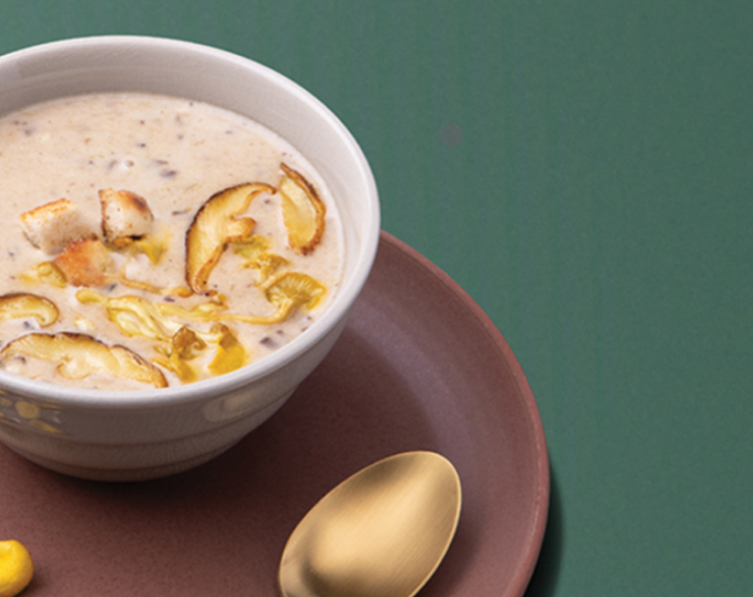White bowl of beige cream of mushroom soup on brown plate with spoon on dark green surface.