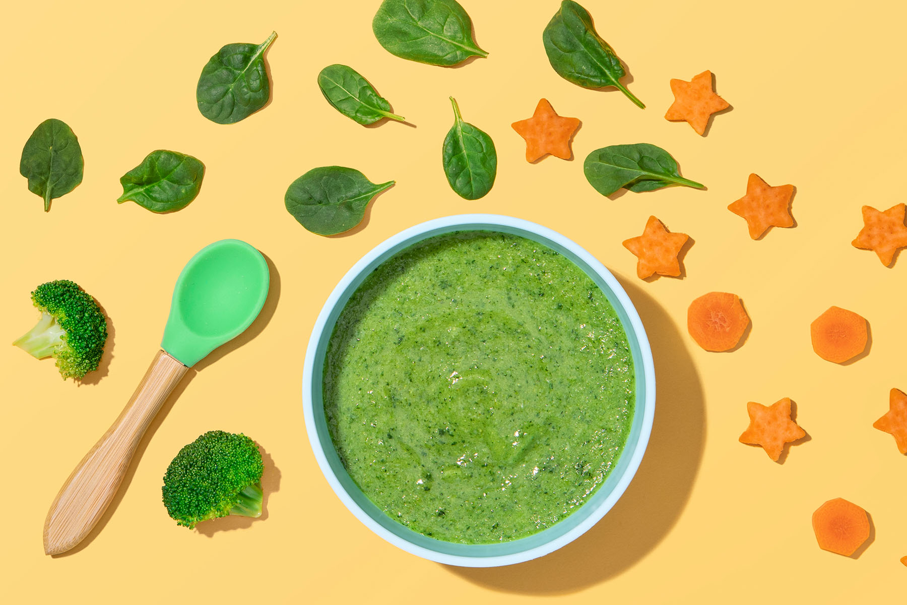 Bowl of blended green veggies with spoon and carrot, broccoli and spinach surrounding