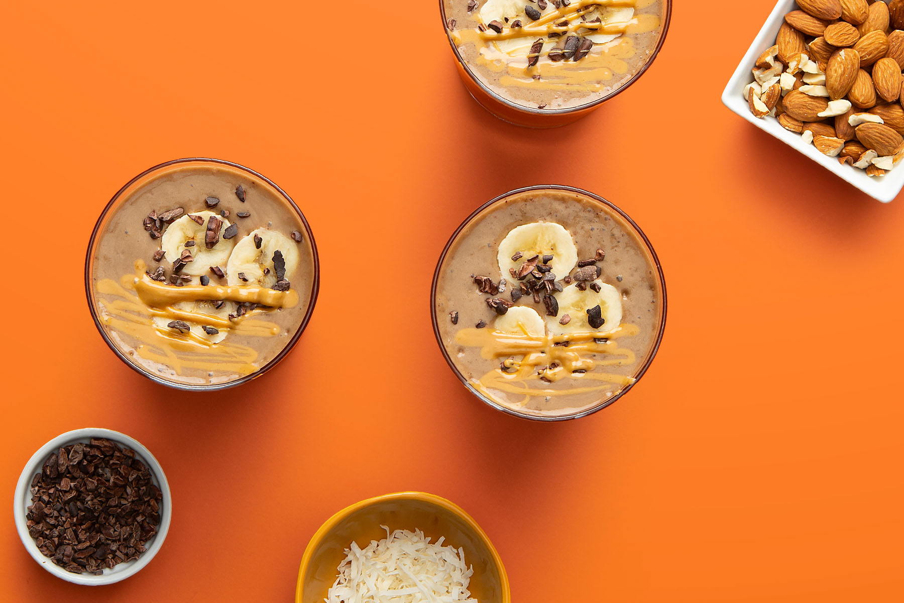 chocolate almond banana smoothie in glasses on orange background with coconut, chopped almonds and cacao nibs
