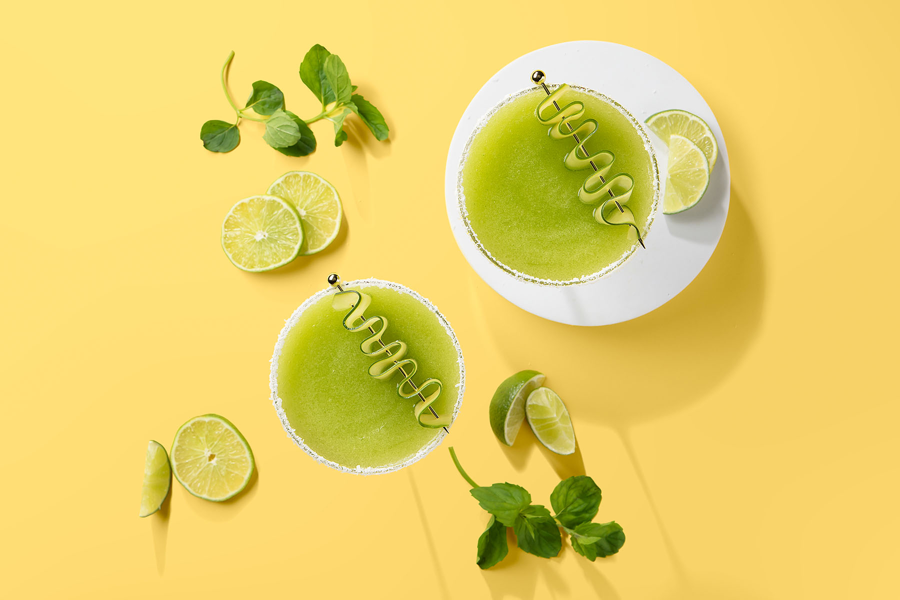 Green cocktail garnished with herbs, fresh lime slices, and cucumber with yellow background