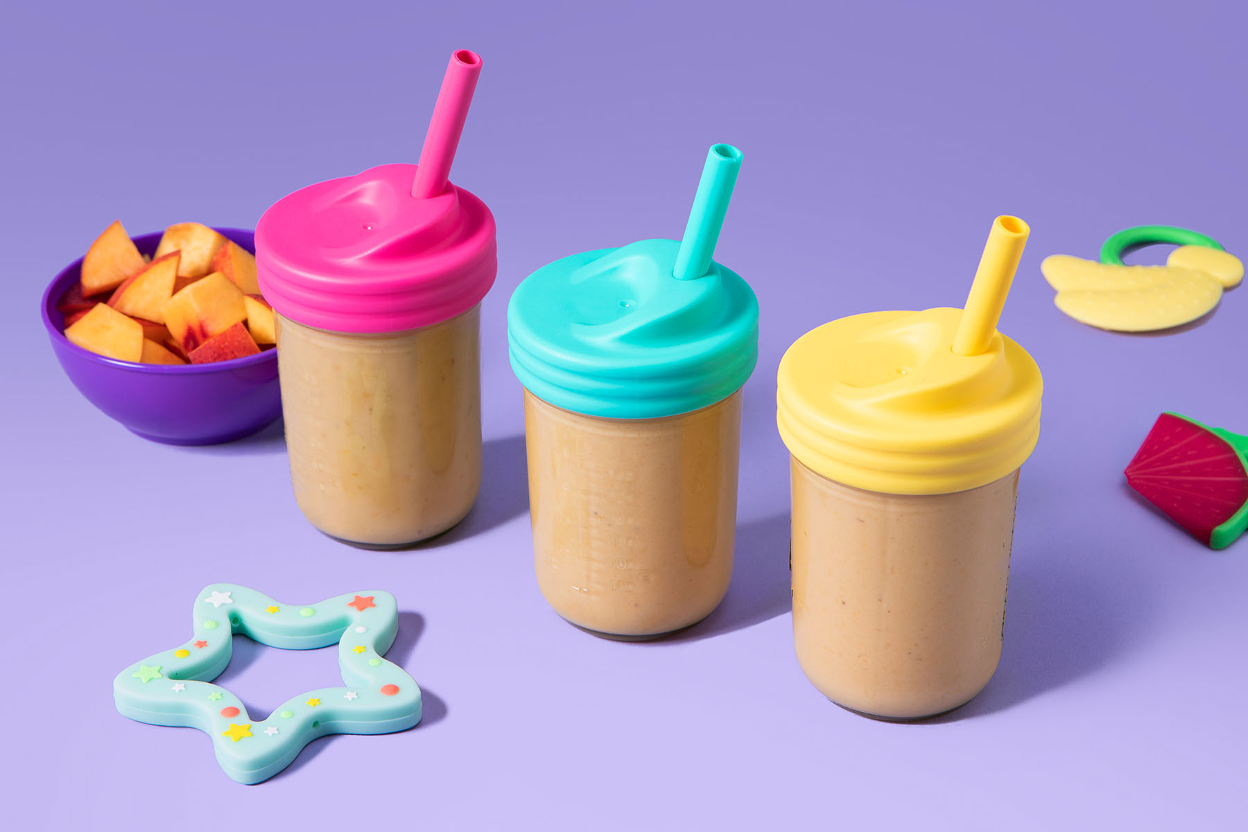 Three peach smoothies in reusable cups with a bowl of cut up peaches and toys
