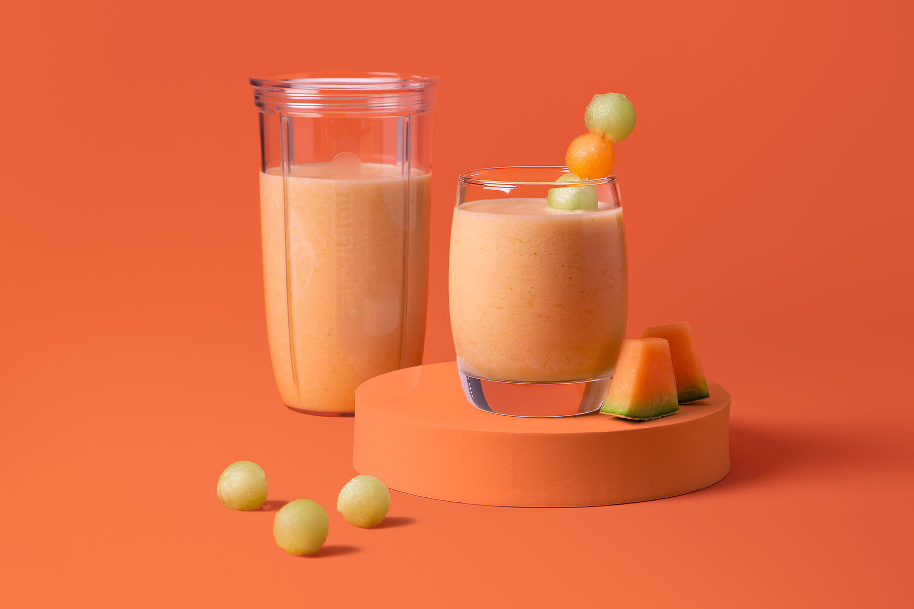 Orange melon cocktail in nutribullet cup and glass garnished with melon ball skewer on orange background