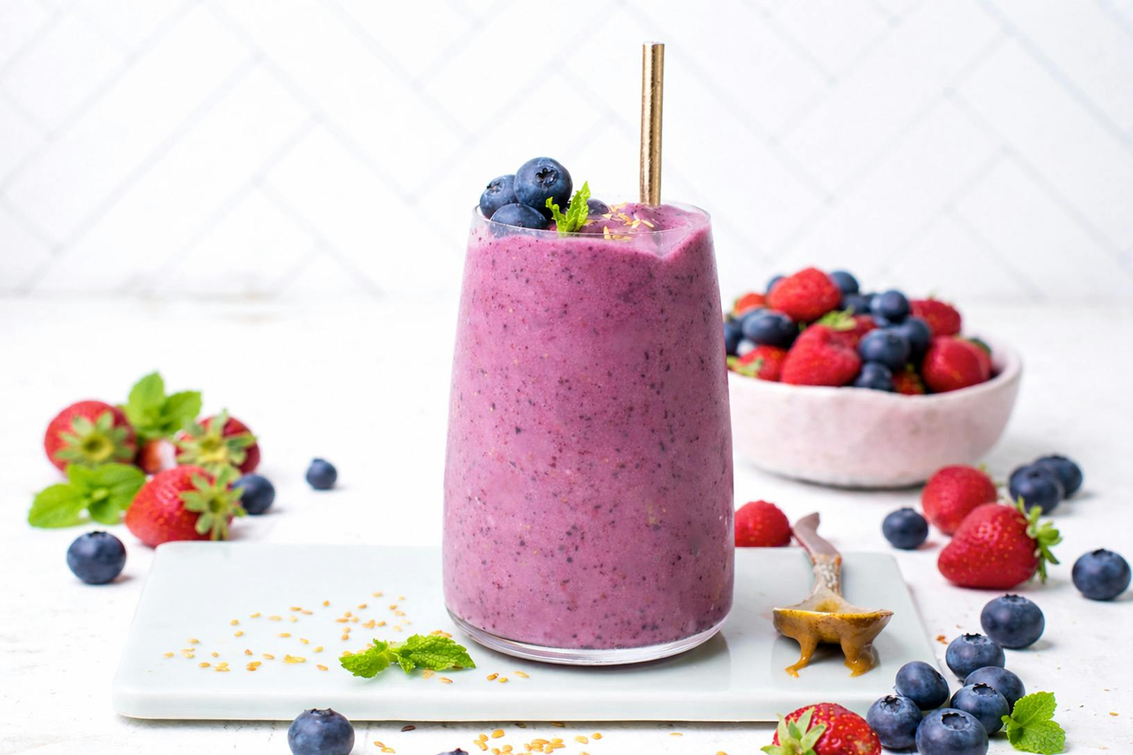 A glass filled with a berry-colored smoothie on a countertop surrounded by berries.