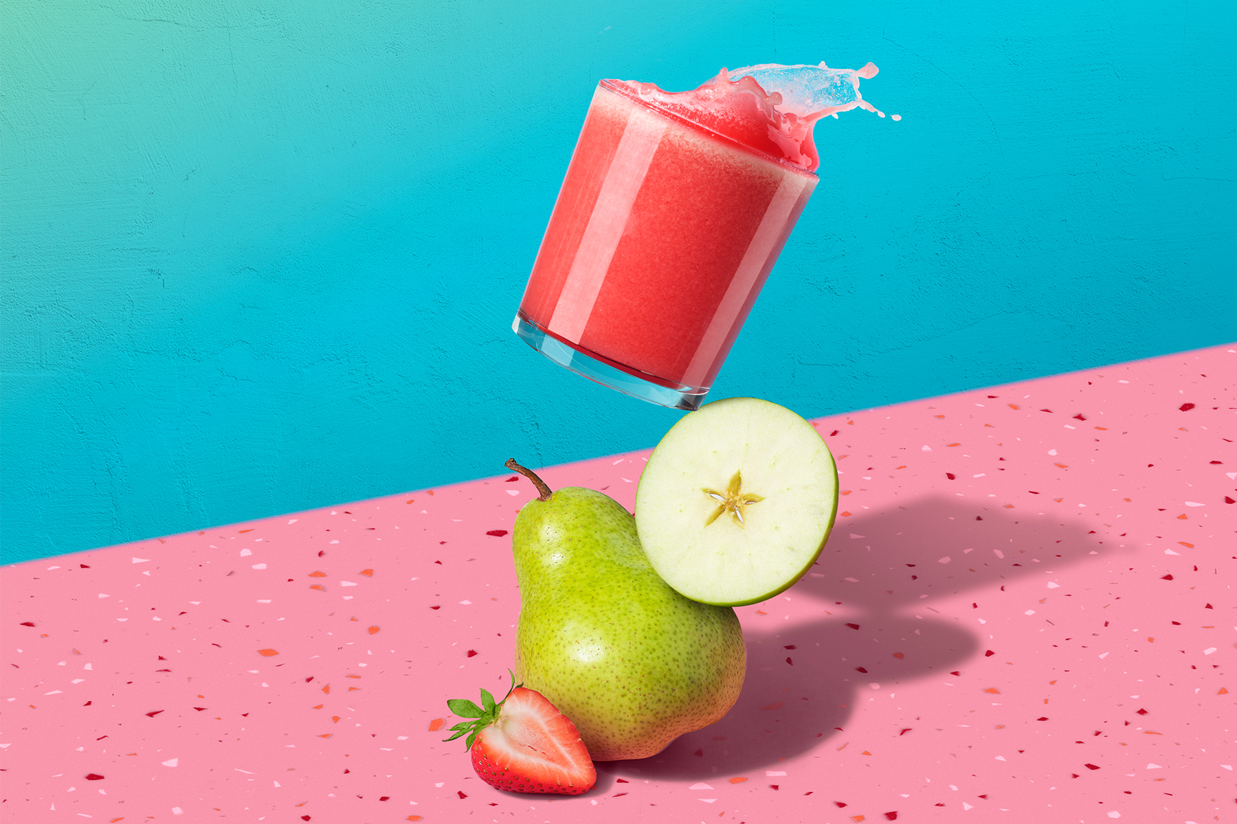 tilted glass sitting on top of pear with strawberry, apple, pear juice splashing on a pink counter with blue background