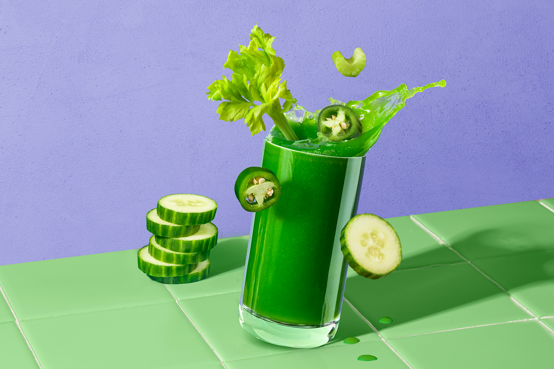 glass of celery cucumber jalapeno juice sitting on a green counter with slices of jalapeno, cucumber and celery splashing out in front of a purple background