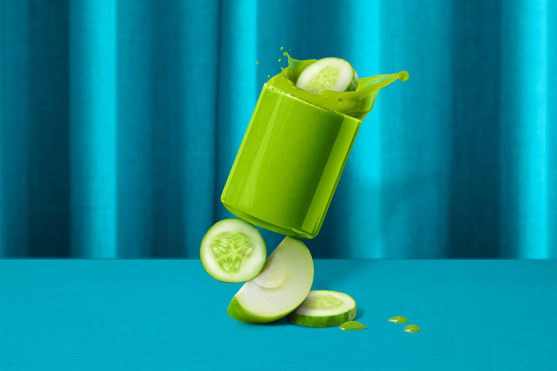 glass of cucumber apple juice sitting on top of sliced cucumber and apple in front of a blue background