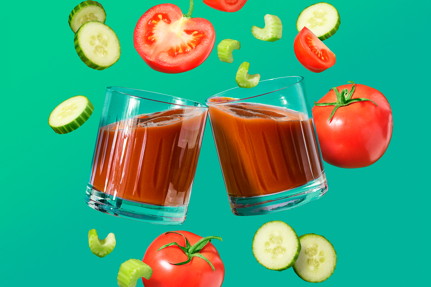 two glasses of tomato juice with tomatoes, cucumber and celery floating in front of green background
